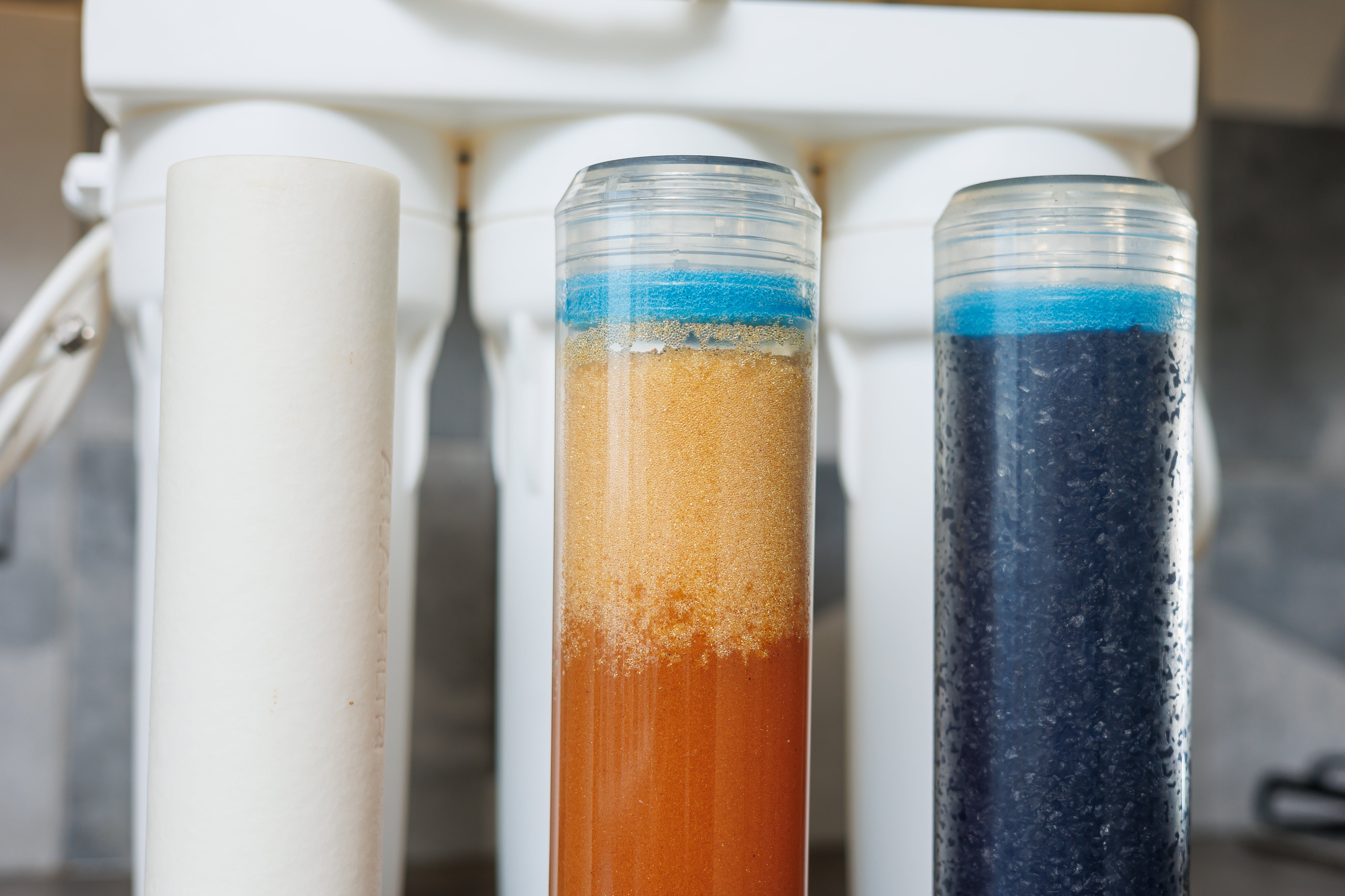 Close-up of a water filter system with two transparent test tubes showing colored sediment layers, set against a white filter housing.