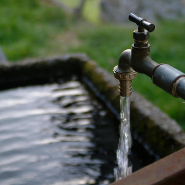 A close-up outdoor shot of a metal faucet running water into a stone basin, with blurred green grass and rocks in the background.