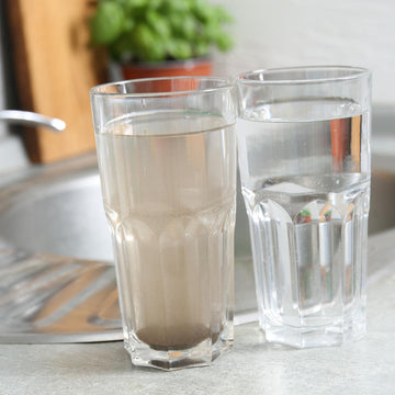 Two clear glass tumblers on a kitchen counter, one filled with cloudy tap water and one with clear water, with a sink and faucet in the background.