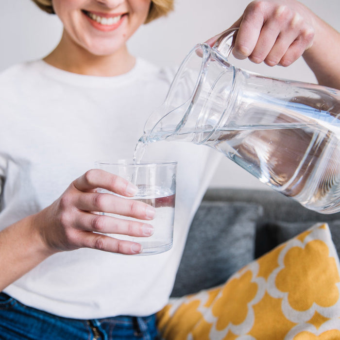 A smiling woman in a white t-shirt pours clean water from a clear glass pitcher into a clear glass tumbler.