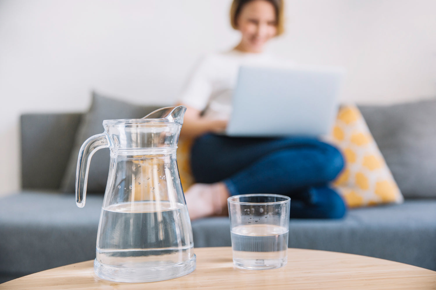 A clear glass water pitcher and matching glass on a wooden table in the foreground, with a blurred woman sitting on a gray sofa using a laptop in the background.