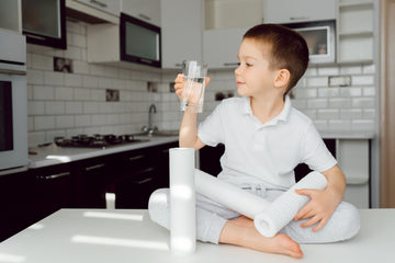 A young boy in a white polo shirt and light gray sweatpants sits cross-legged on a white kitchen counter, holding a clear glass of water. 