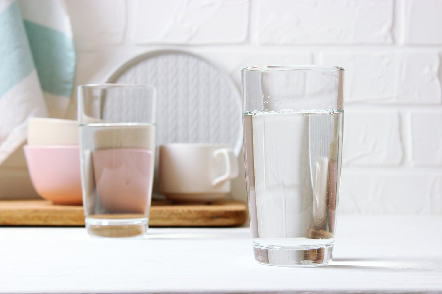 Two clear glass drinking cups on a white tabletop, with a softly blurred kitchen or dining background including pastel bowls, a mug, and a white brick wall.