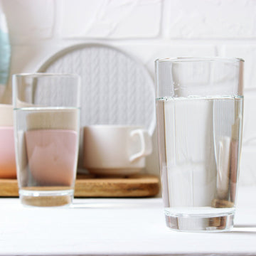 Two clear glass drinking cups on a white tabletop, with a softly blurred kitchen or dining background including pastel bowls, a mug, and a white brick wall.