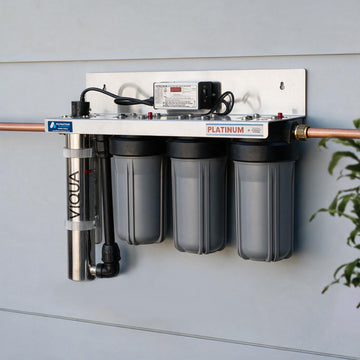 A wall-mounted water filtration system with three gray filter canisters, a copper pipe connection, a vertical stainless steel pre-filter, and a power supply on a white backplate against a light gray wall.