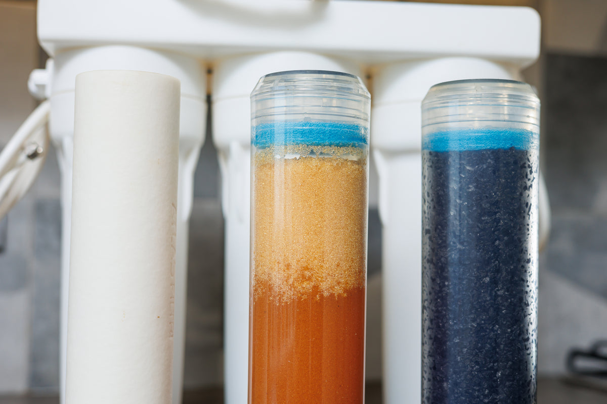 Close-up of a water filter system with two transparent test tubes showing colored sediment layers, set against a white filter housing.
